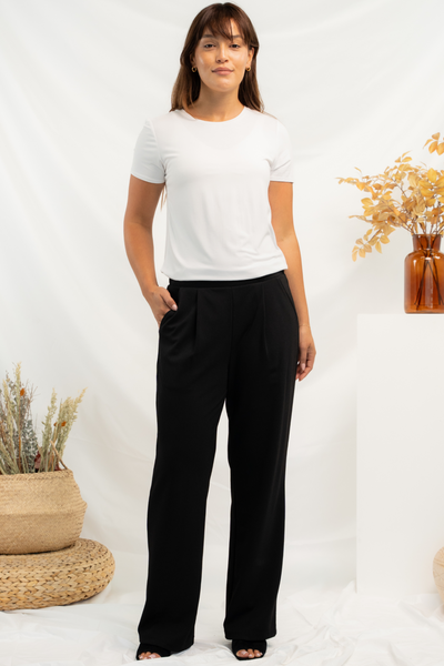 Woman wearing a white t-shirt and black wide leg pants standing against a white background with decorative fall plants.