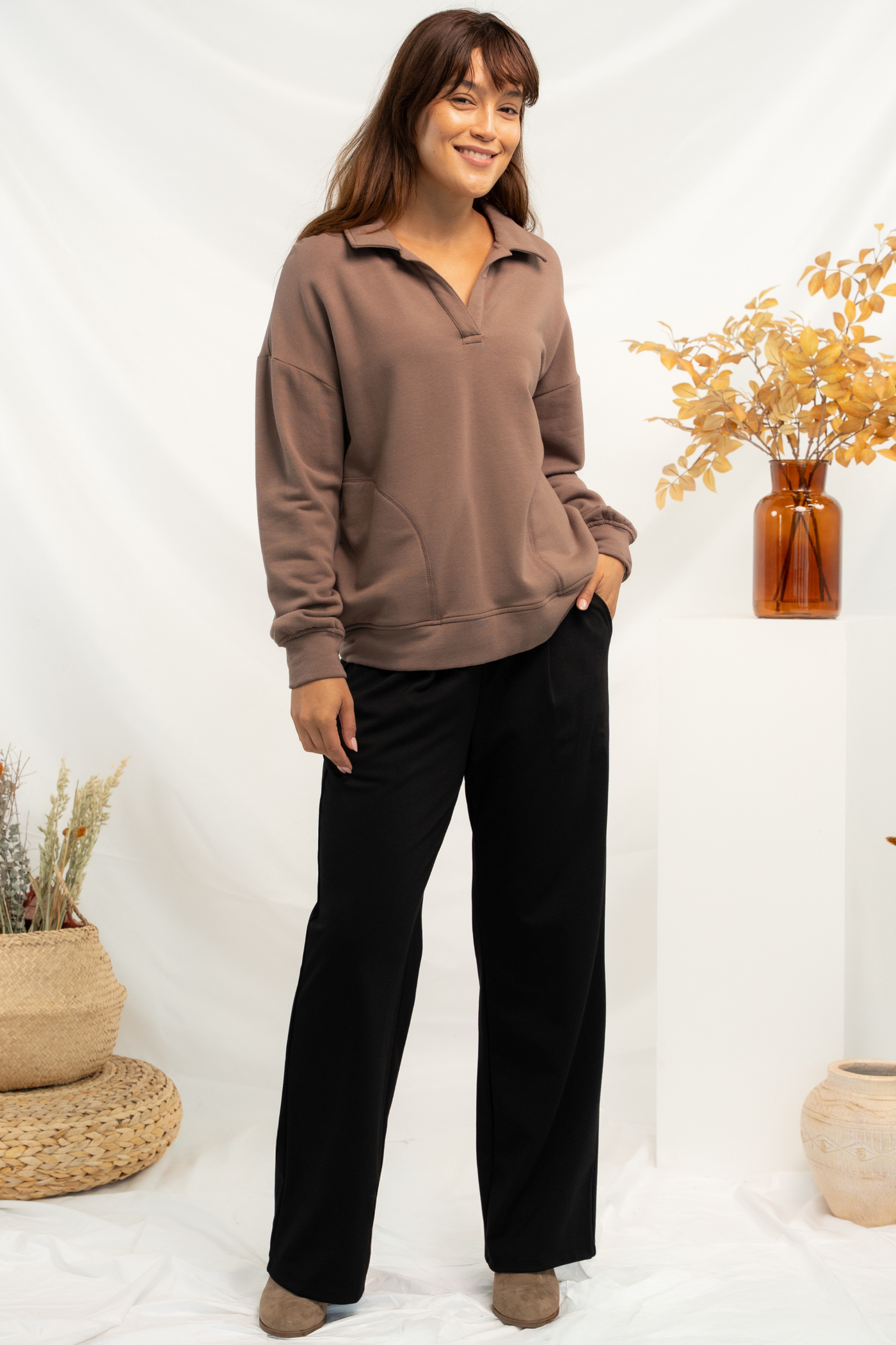 Woman wearing a brown collared sweatshirt and black pleated pants with brown heeled boots, standing against a white background with decorative fall elements.