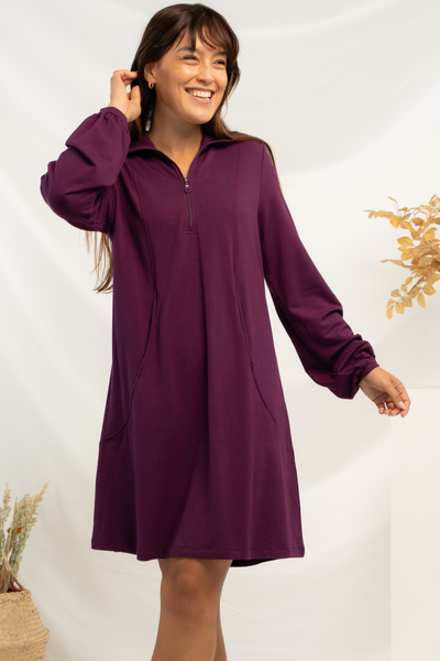 A woman wearing a purple a-line long sleeve, knee length dress with quarter zip detail, paired with black boots.