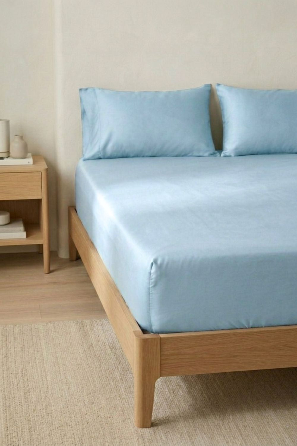 Bed with light blue bamboo fitted sheet bedding and pillows in a room with wooden furniture and neutral walls.