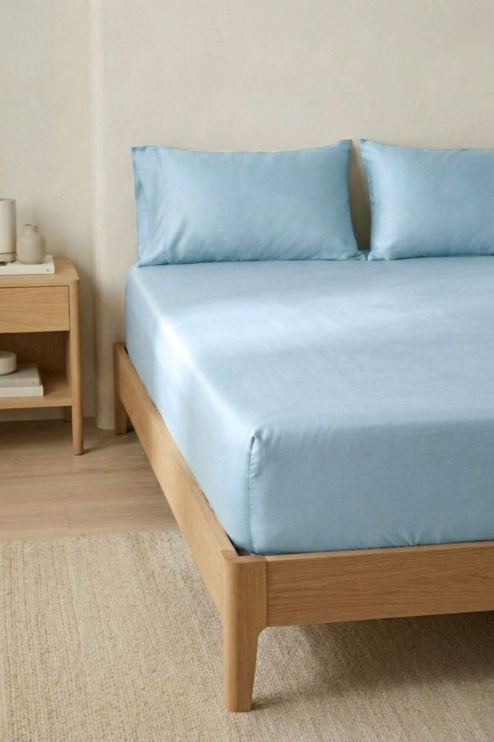 Bed with light blue bamboo fitted sheet bedding and pillows in a room with wooden furniture and neutral walls.