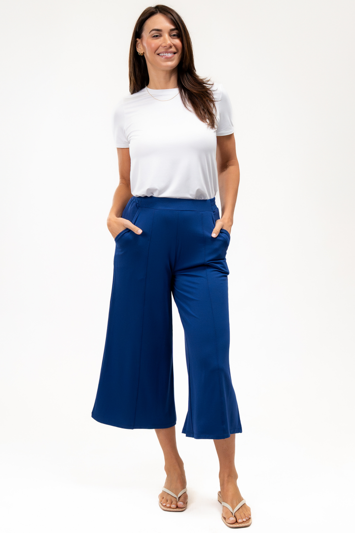 A woman wearing a white t-shirt and blue culotte pants with white sandal flip flops on a white background