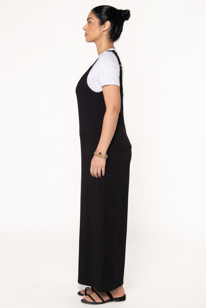 Model wearing a bamboo jumpsuit in Black which features wide legs, pockets, and adjustable thin straps. Layered underneath the jumpsuit is a white t-shirt and paired with black sandals. 
