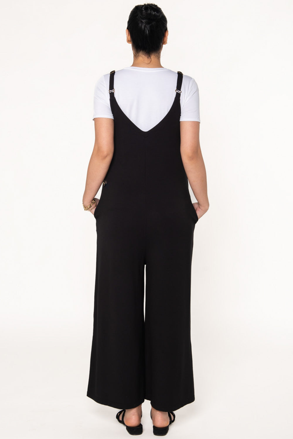 Model wearing a black wide leg jumpsuit with pockets and adjustable thin straps. Layered underneath the jumpsuit is a white t-shirt. 