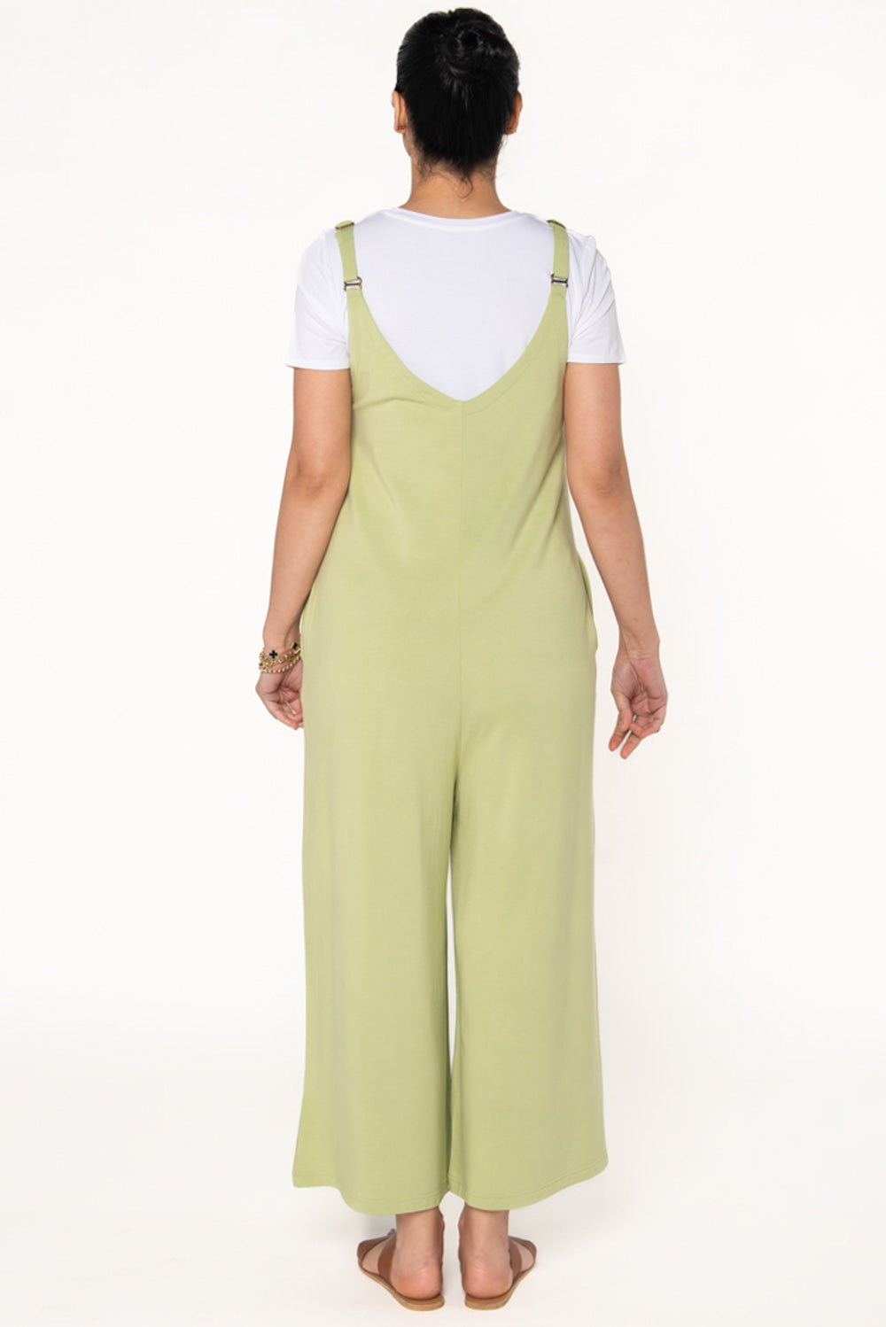 Model wearing a Citron green bamboo jumpsuit with wide legs, pockets, and adjustable thin straps. Layered underneath the jumpsuit is a white t-shirt. Model is showing the back of the jumpsuit.