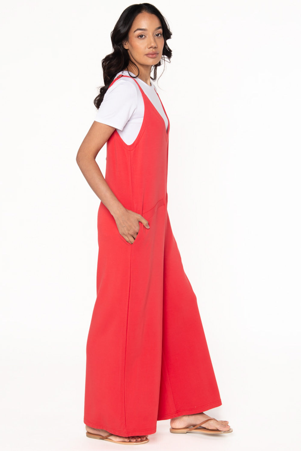 Model wearing a Melon coloured bamboo jumpsuit with wide legs, pockets, and adjustable thin straps. Layered underneath the jumpsuit is a white t-shirt. Model is showing the side view of the jumpsuit.